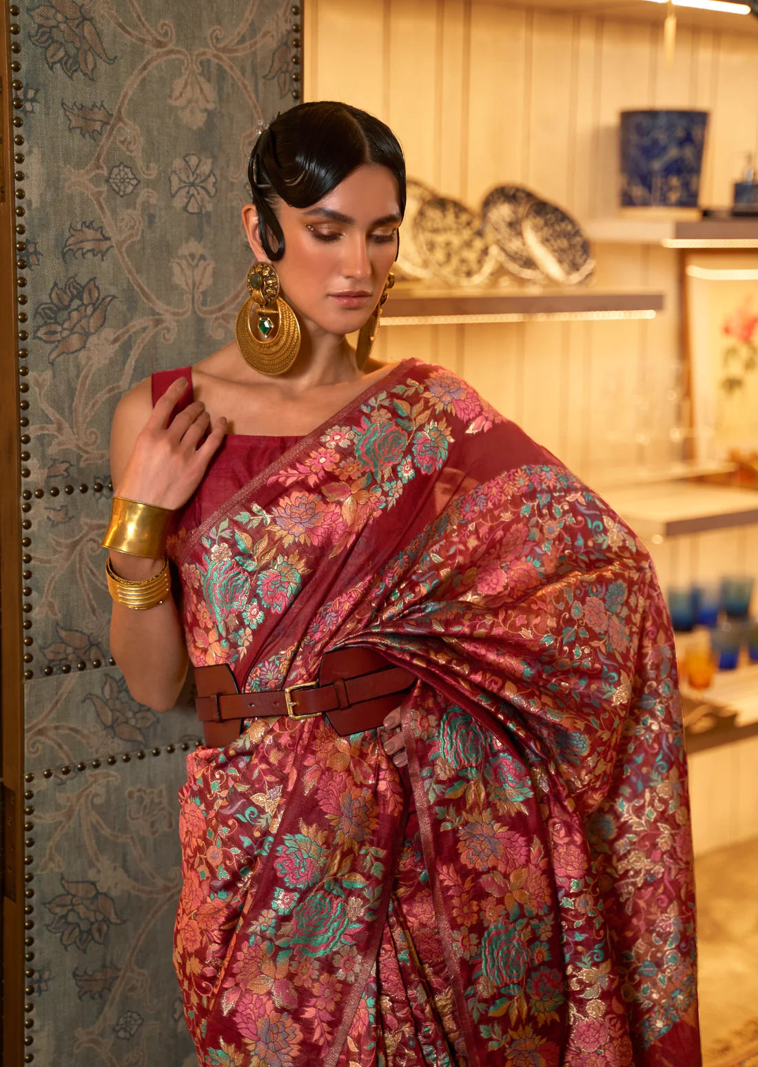 woman in maroon red kashmiri silk pashmina saree wearing gold earrings & bangles along with a brown leather belt.