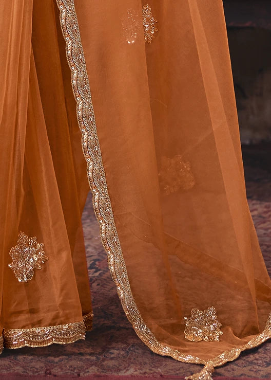 Close-up of a rust-orange traditional organza saree pallu with metallic gold embroidered borders and floral embellishments, falling adjacent to saree pleats.