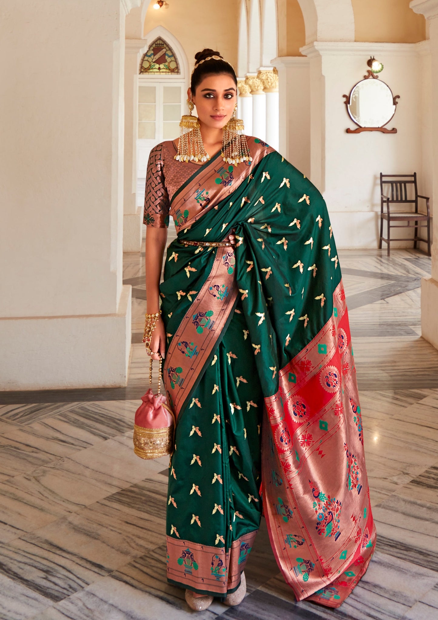 Woman posing in daark green bridal paithani saree with heavy gold zari pallu holing pink potli bag in right hand.
