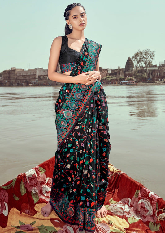 A woman standing on a boat wearing a traditional Kashmiri pashmina silk embroidered black handloom saree with a floral jamawar motif and a red and green border.