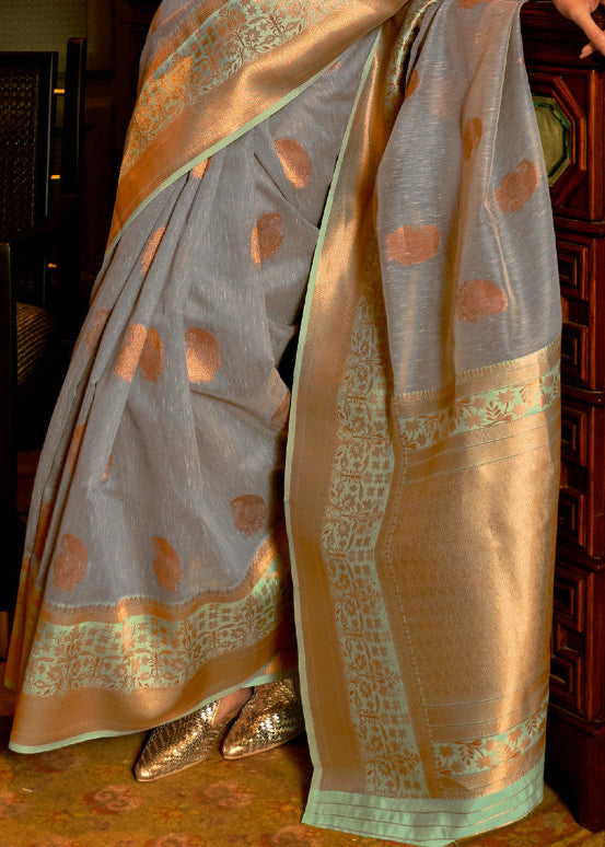 Close-up of a draped saree in a light gray-blue linen silk with metallic gold circular motifs, paired with a wide gold border and mint green trim, shown on a model in a traditional setting.