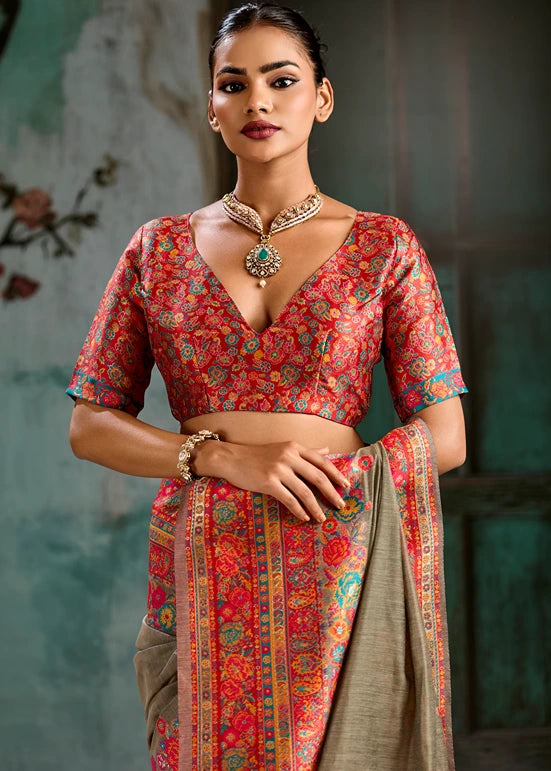 A woman modeling a traditional Indian outfit featuring a red and orange floral printed blouse with a deep V-neckline and short sleeves, paired with a matching draped saree in a muted olive-beige color with a decorative border.