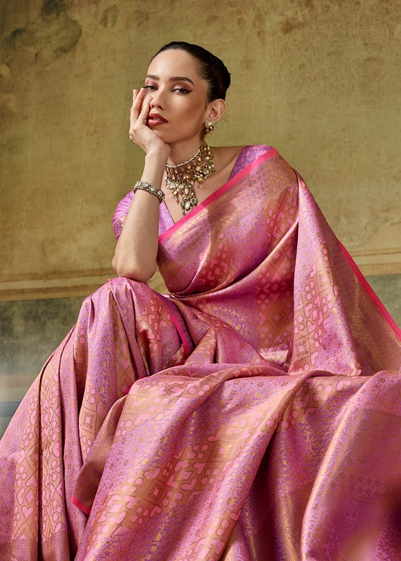 A woman wearing a baby pink and gold patterned kanchipuram silk saree, seated against a textured beige wall, wearing matching pink blouse and gold jewelry.