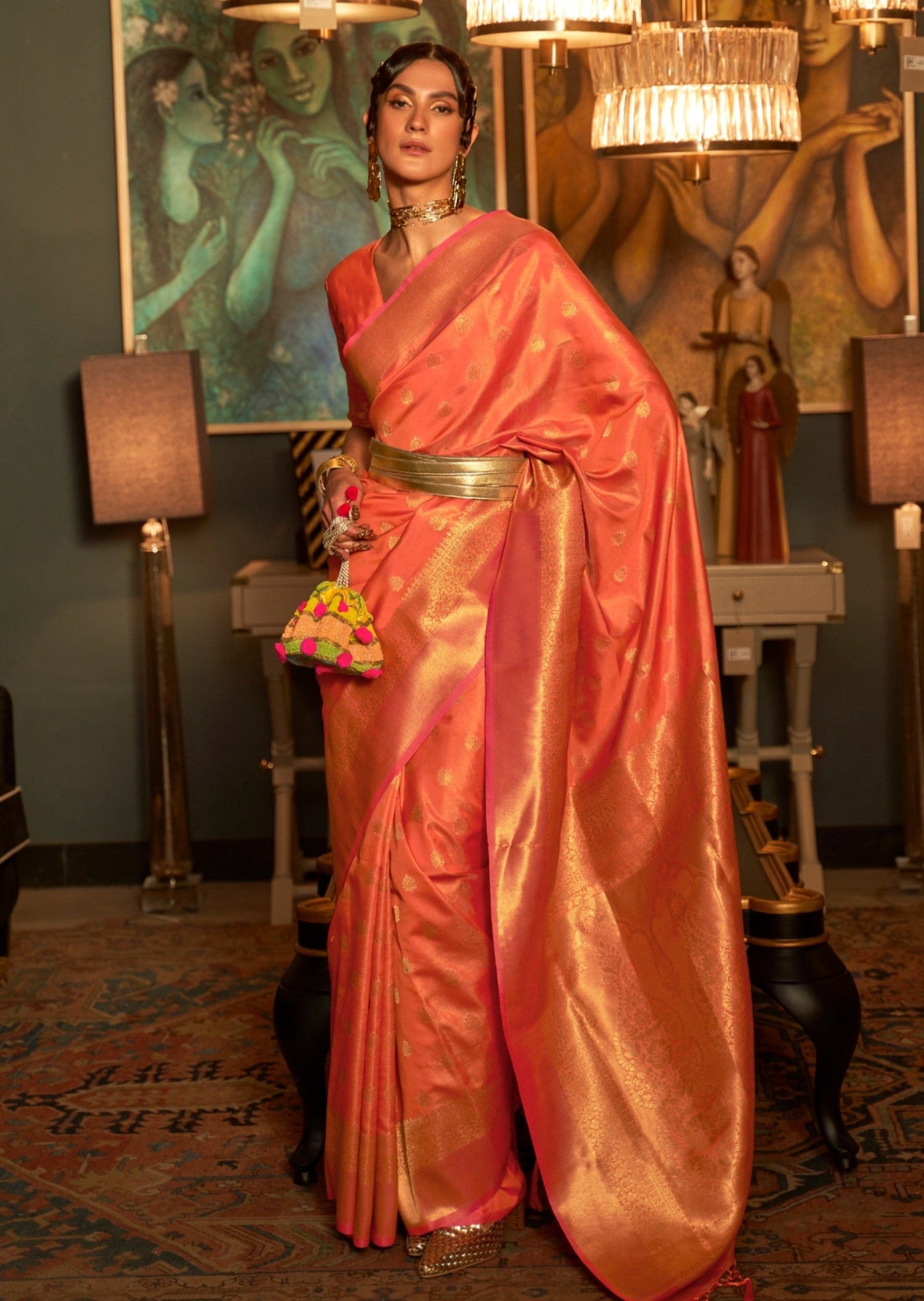 woman standing in orange colour banarasi silk saree below glowing golden chandelier