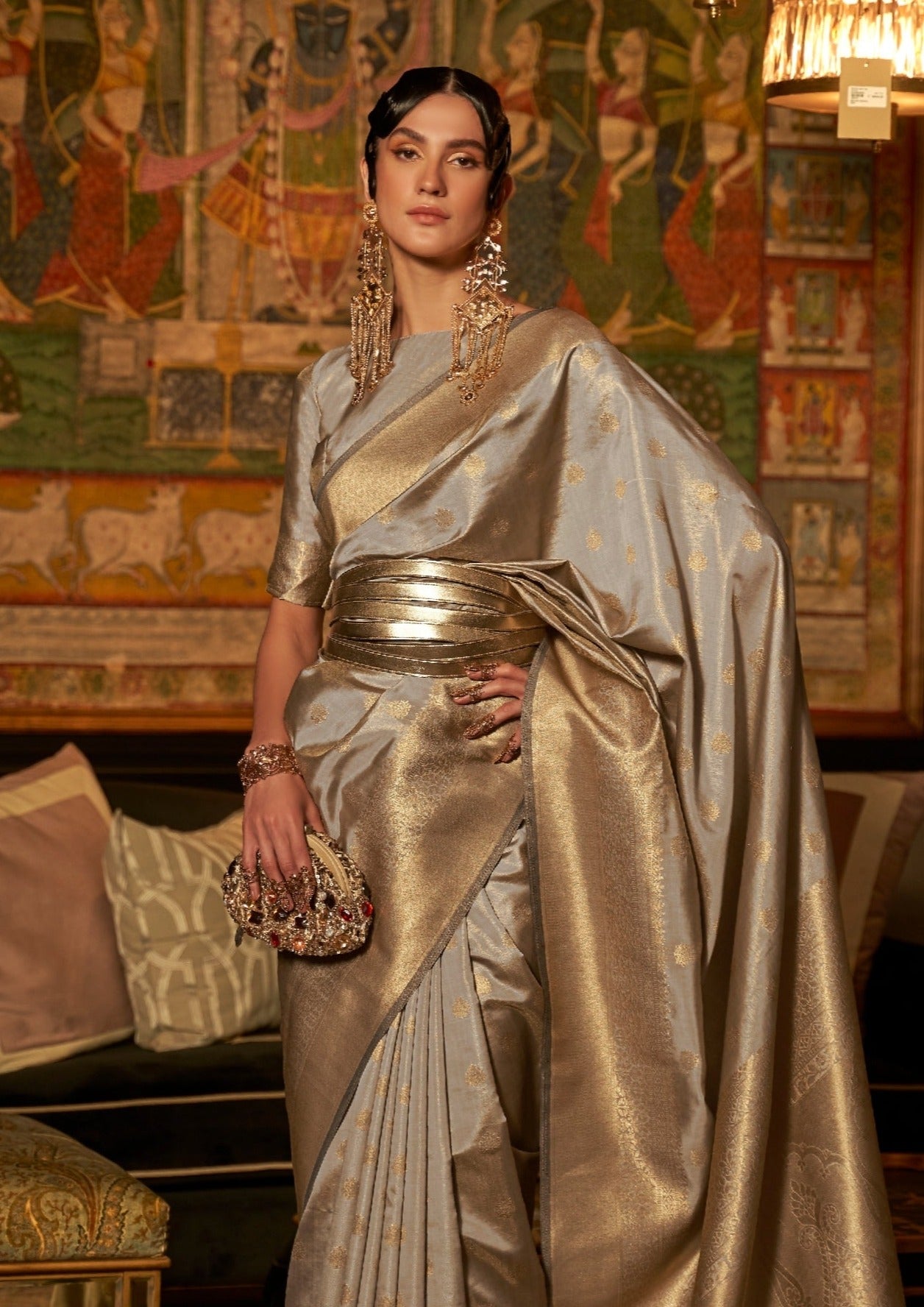 woman standing in front of black sofa wearing banarasi Silk silver Handloom Bridal Saree & golden belt holding golden purse in hand