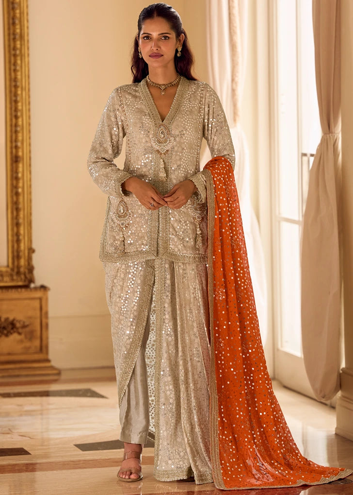 Woman in a sparkling beige jacket dhoti indian outfit with an orange dupatta with heavy sequins embroidery work.