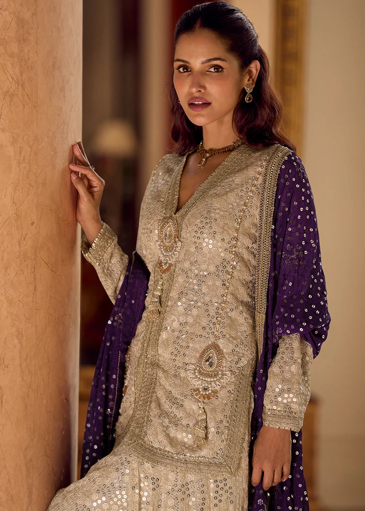 A woman wearing an ornate beige embellished traditional outfit in usa with a deep V-neck, long sleeves, and a purple dupatta draped over her shoulders, posed against a warm-toned indoor wall.