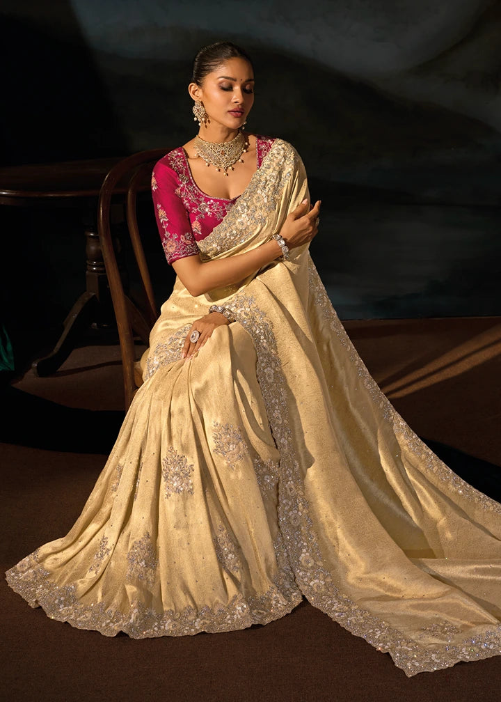 A woman seated on a dark wooden chair wearing a flowing luxury cream kanjivaram silk saree with peach floral embroidery and a bright red embroidered designer blouse, photographed in a dim indoor setting.