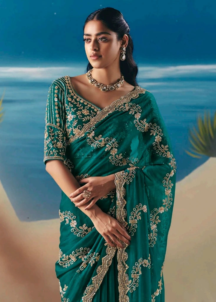 Woman in an emerald green saree with gold beadwork and sequins embroidery against a blue ocean background.