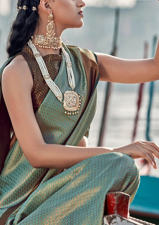 A woman wearing a sage green kanjivaram silk saree with gold zari embroidery, a black sleeveless blouse, and ornate gold jewelry, posed outdoors.