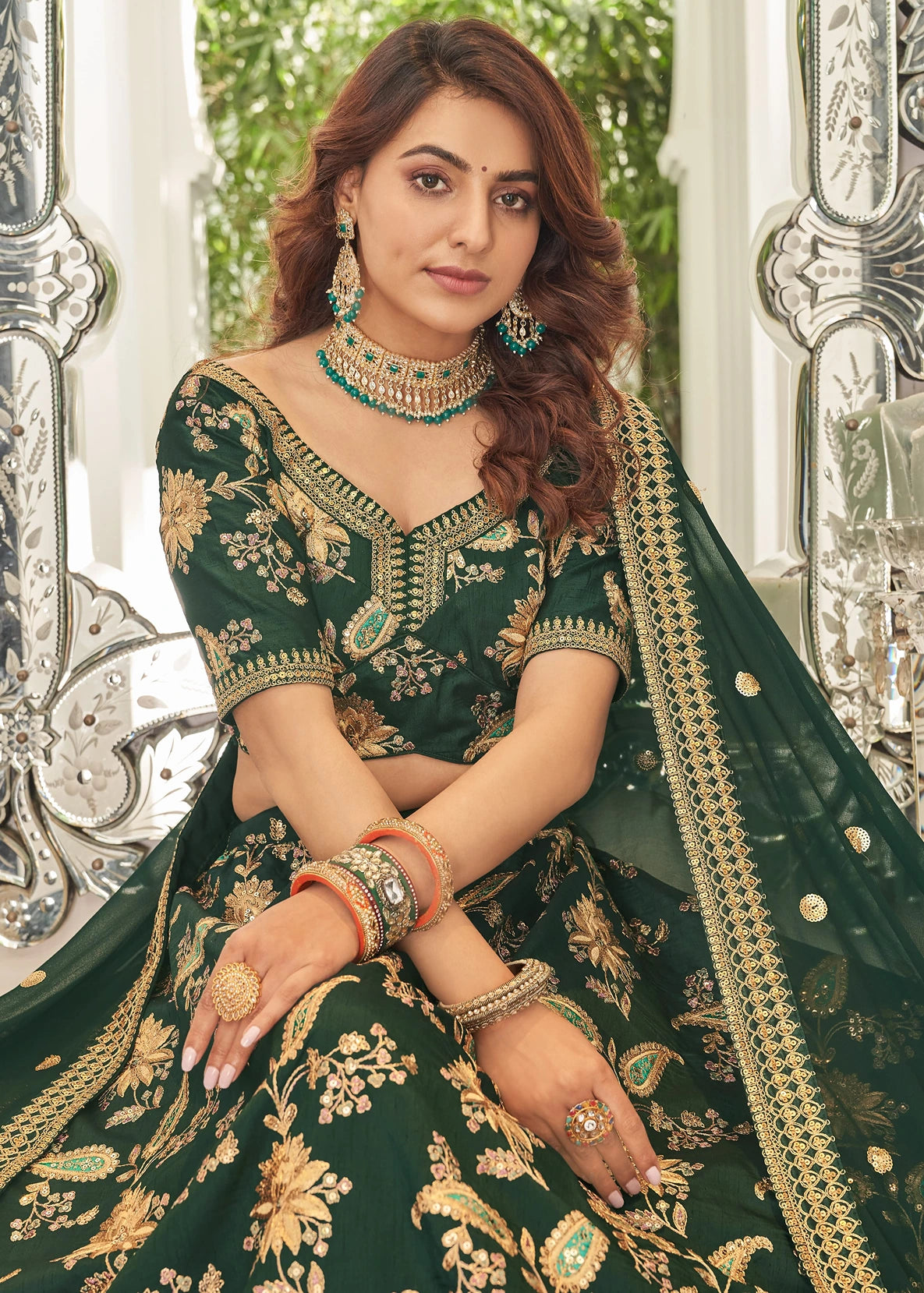 Woman in a green and gold traditional outfit with jewelry, sitting against an ornate silver and white background; showcasing latest lehenga collection in Malaysia online.