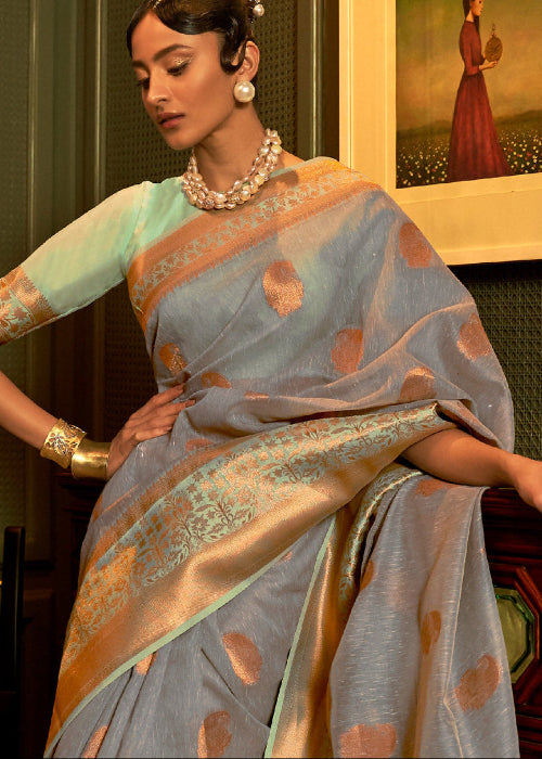 A woman wearing a grey handloom linen silk saree with gold zari motifs, paired with a mint green blouse, posing in an indoor setting with a framed painting in the background.
