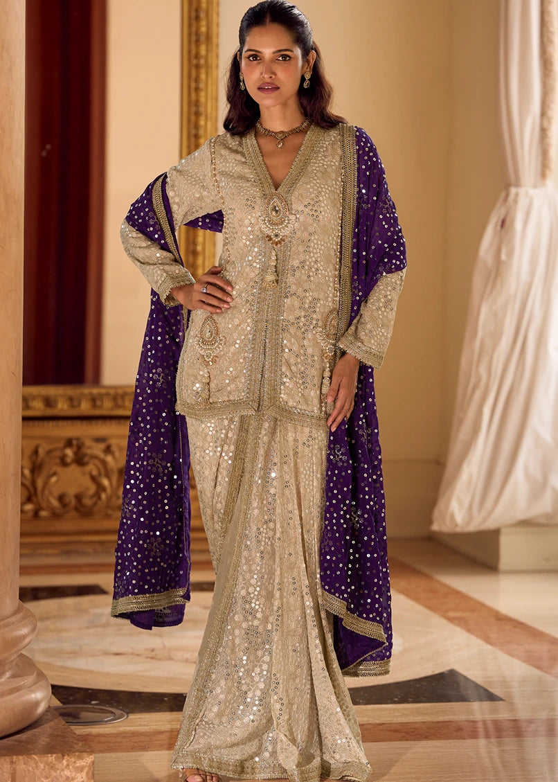 A woman models a traditional Indo western outfit set consisting of a heavily embellished beige kurta and matching dhoti skirt in usa with a deep purple dupatta draped over her shoulders, set against an elegant indoor background.