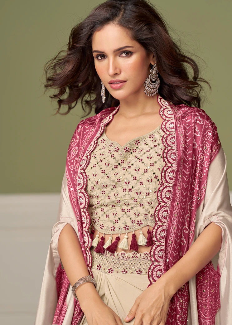 Traditional south asian indo-western outfit featuring a beige embroidered blouse with tassel details, a matching beige dhoti skirt, and a prominent pink bandhani shrug style jacket over her shoulders, listed for free delivery to USA and worldwide.