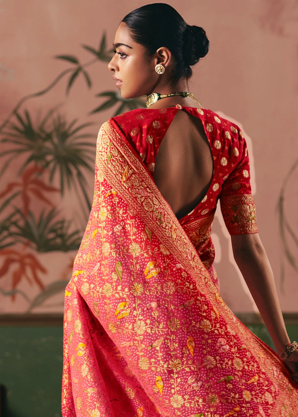 Back view of a woman in a light pink bandhani saree and red blouse with floral & bird patterns, showcasing pallu details.