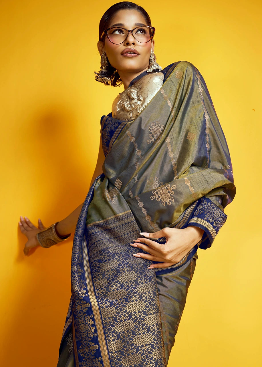Indian bride in a traditional Indian kanjivaram silk saree against a solid yellow background. The saree is a muted olive green with intricate gold and blue floral zari patterns, paired with a matching navy blue blouse.