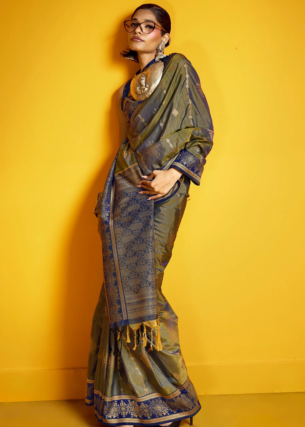 A woman wearing a metallic olive green and navy blue patterned saree with gold zari detailing, styled with a matching dupatta, gold necklaces, and glasses, posing against a solid yellow background.