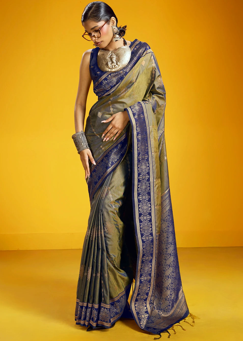 A woman modeling a traditional Indian kanjivaram silk saree with a muted military green body and a deep navy blue border & blouse with intricate gold zari patterns, set against a solid yellow studio background.