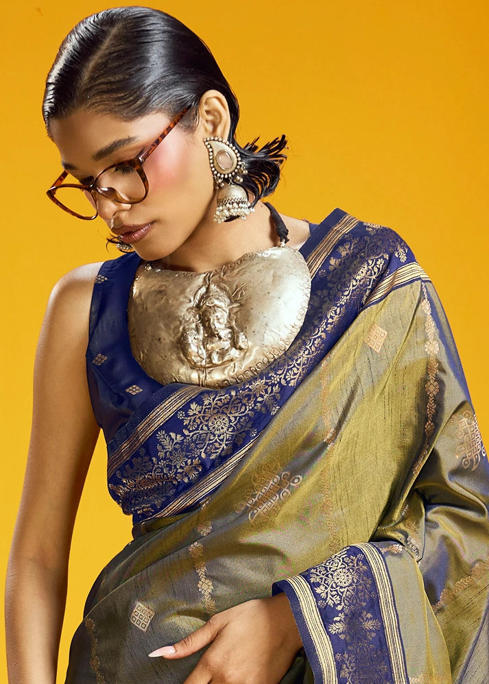 A woman wearing a blue and olive green patterned silk saree with a large metallic gold collar ganesha necklace, styled with traditional earrings, set against a solid yellow background.