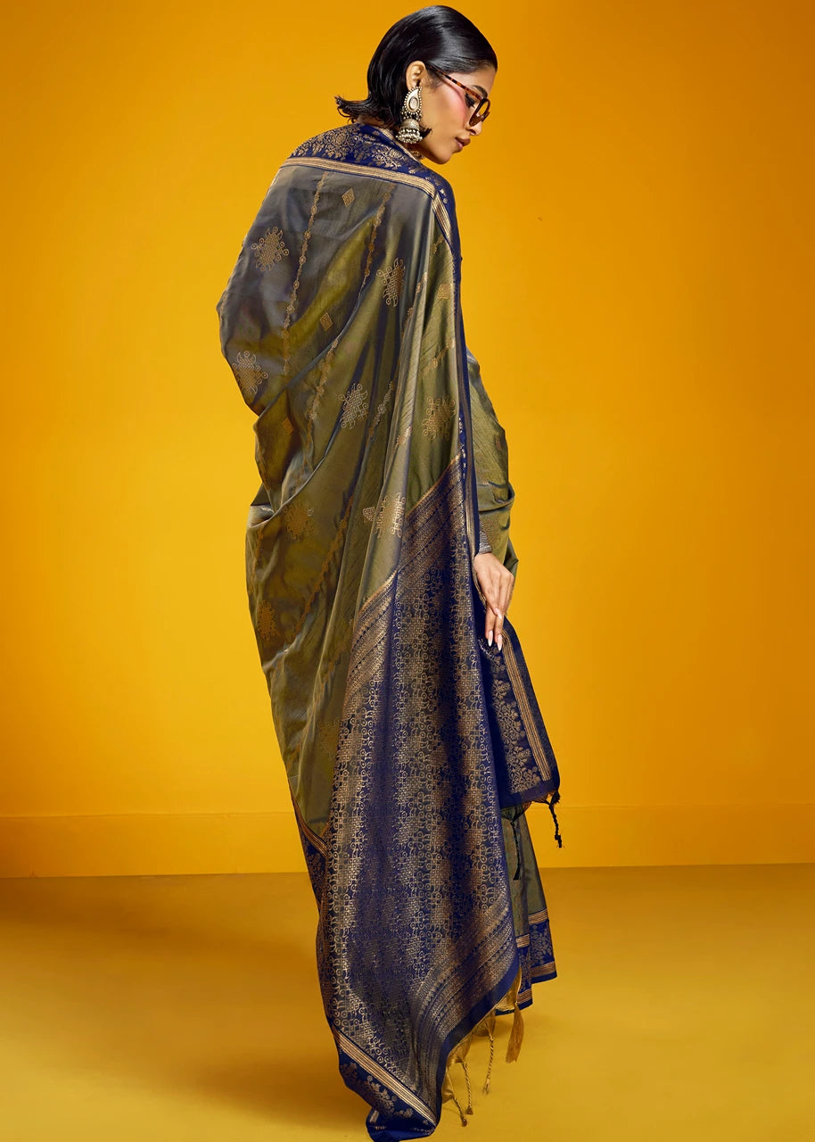 A woman modeling a dark olive green and navy blue patterned saree with gold zari motifs, posed against a solid yellow studio backdrop.