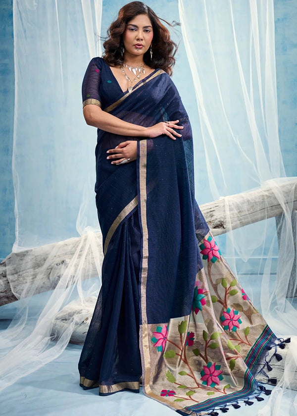 A woman models a dark navy blue muga silk handloom paithani saree with a golden border and a floral printed pallu featuring pink, green, and blue flowers, posed against a light blue backdrop with sheer elegance.