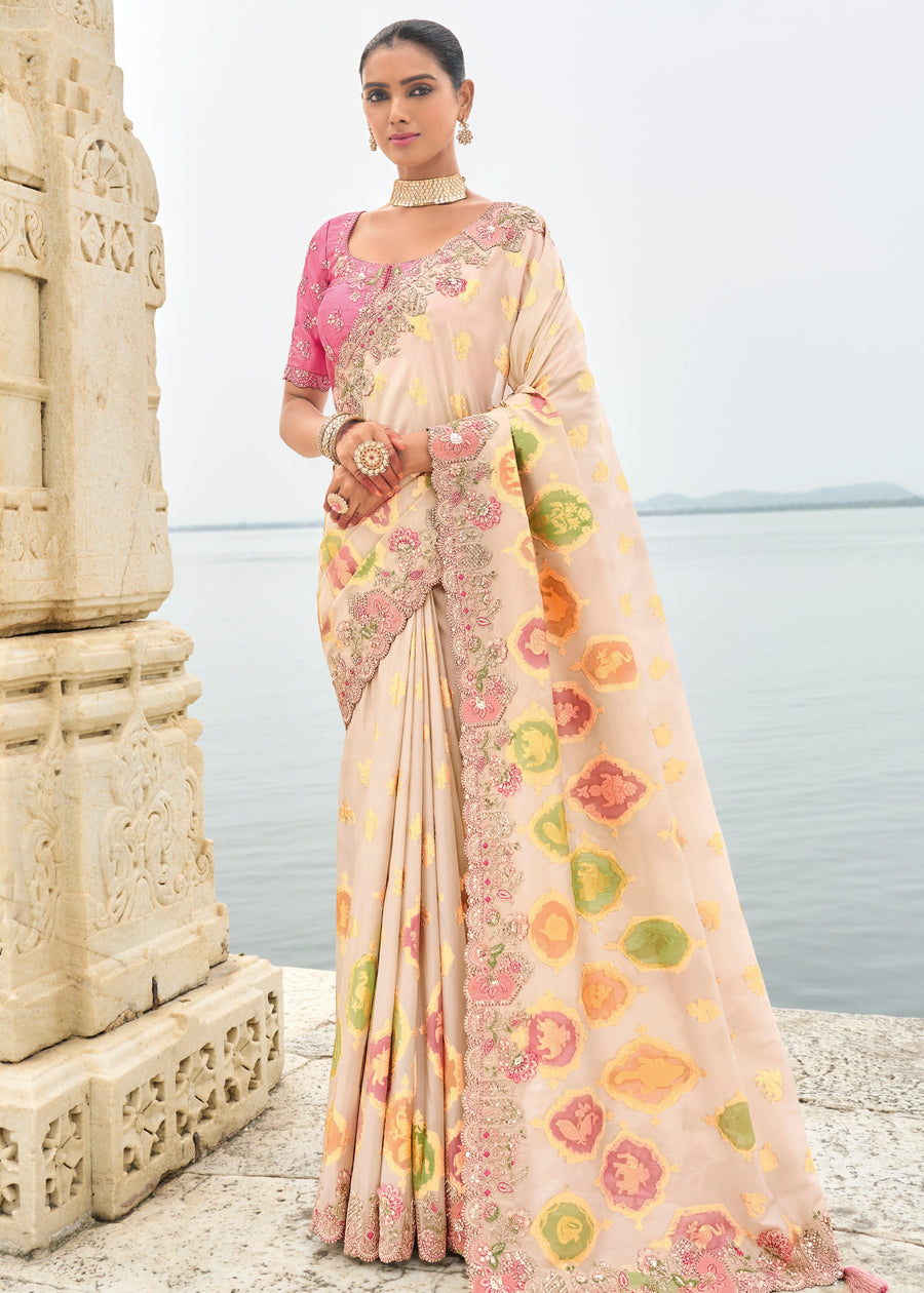 A woman modeling an off-white Banarasi viscose tissue silk saree with embroidery and a cut work border, alongside a pastel pink blouse piece. The saree is displayed in an outdoor setting with ancient architectural elements in the background.