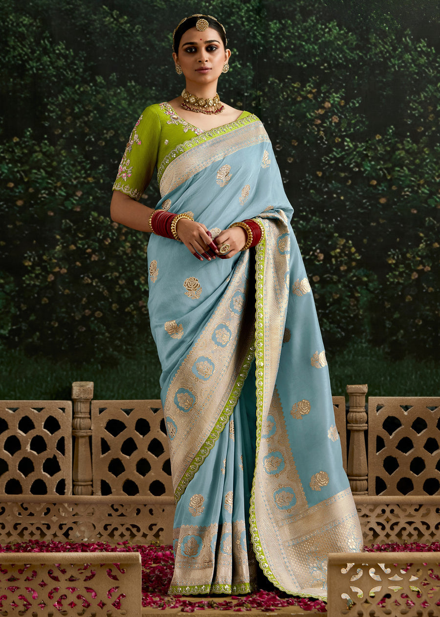 A woman wearing a pastel blue banarasi saree with gold zari rose motifs, a lime green border, and a lime green embroidered blouse, styled with red bangles and gold jewelry against a dark green textured background.