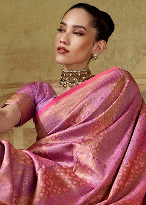 A woman wearing a pastel pink and gold zari woven kanchipuram silk saree with a matching pink blouse, set against a textured wall.