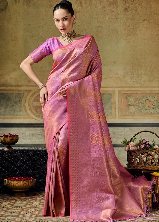 A woman models pastel pink kanchipuram silk saree with gold zari geometric patterns and a matching pink blouse, posed in an ornate room.