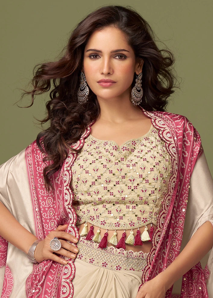Traditional Indian indo-western outfit featuring a beige colored embroidered blouse with pink and gold floral patterns and mirror work, a matching dhoti skirt, and a pink bandhani embroidered shrug jacket USA, set against a solid olive green background.
