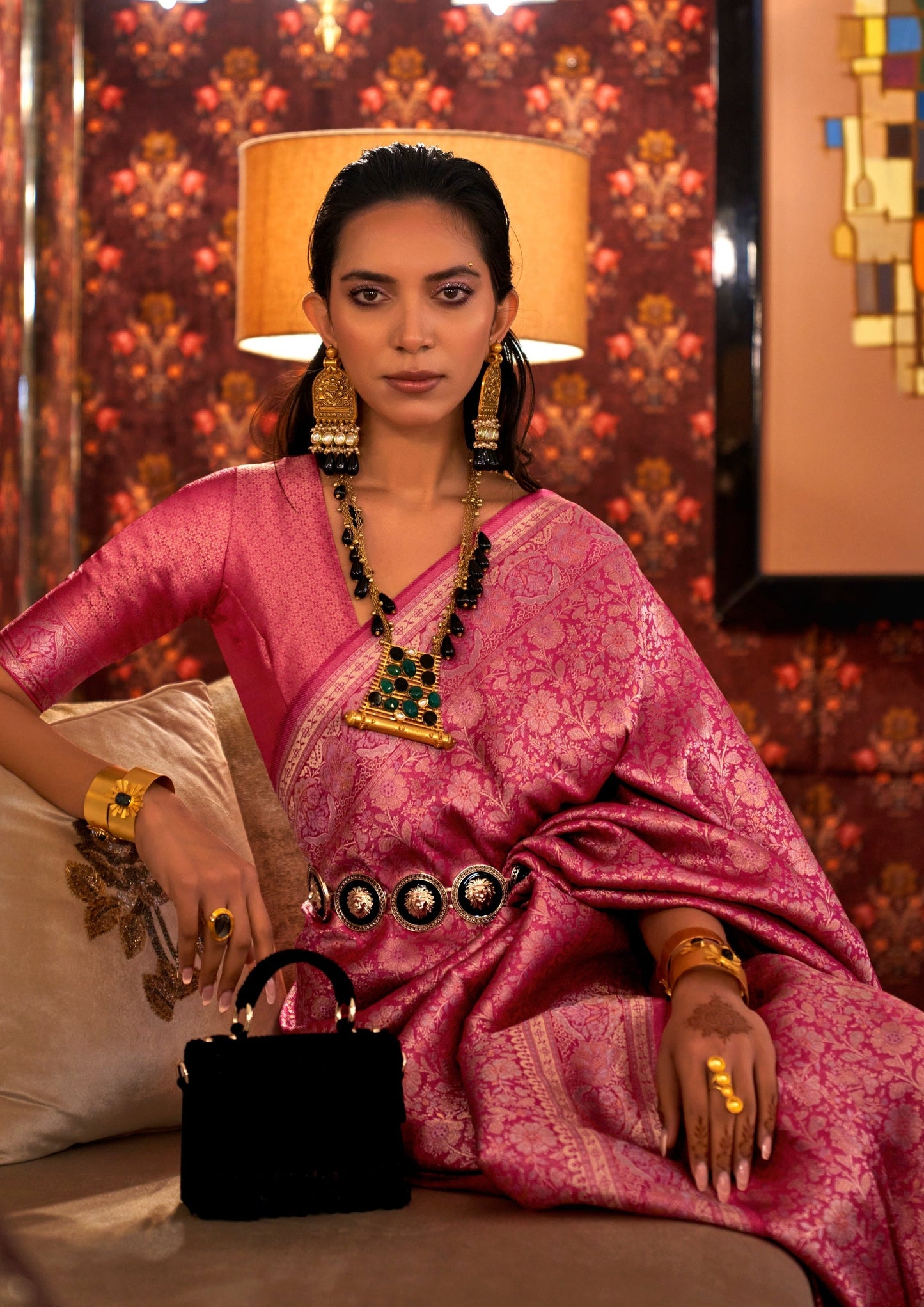 woman sitting in white sofa wearing pink banarasi silk saree and gold jewelry in front of a wallpaper wall having painting hung