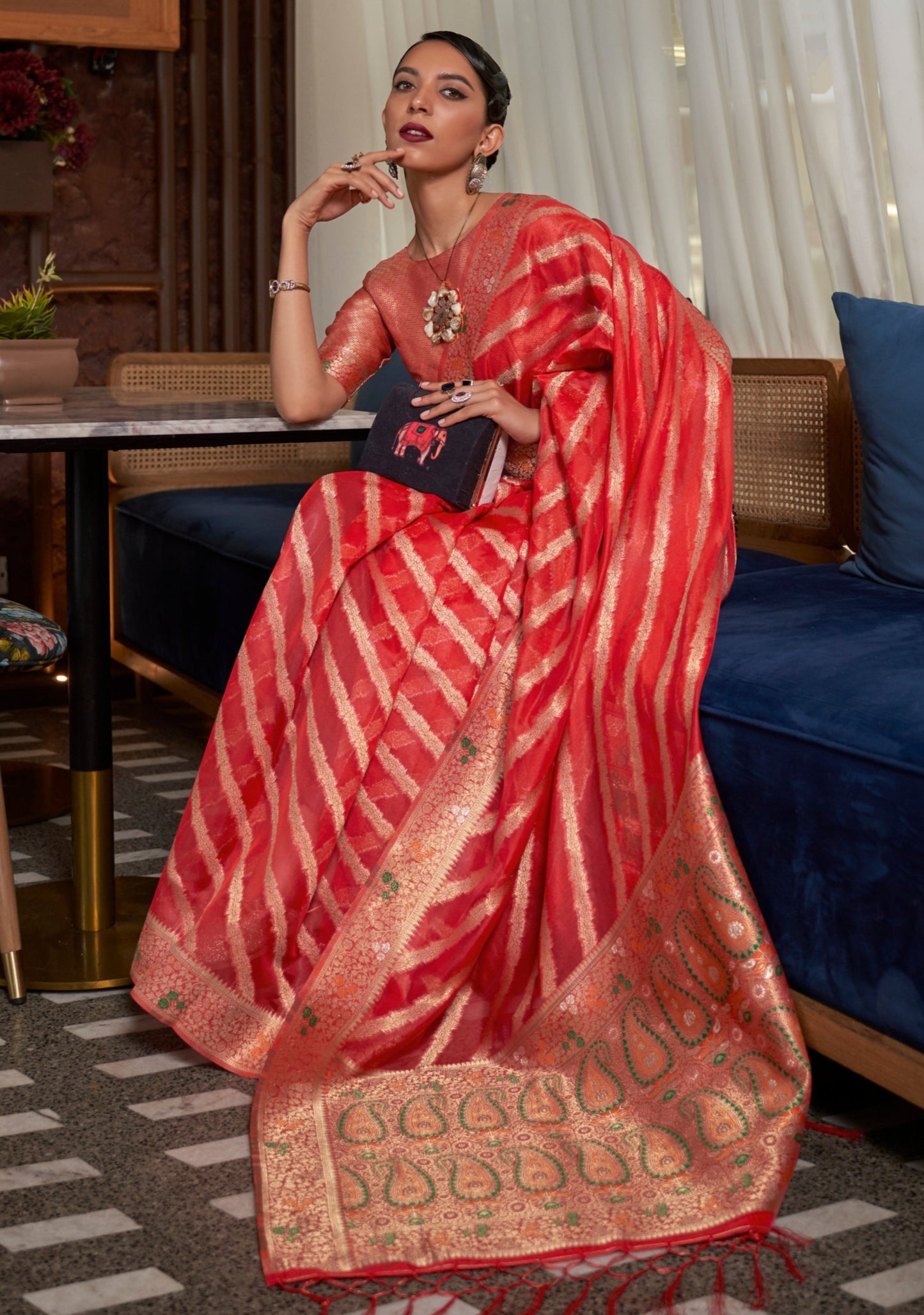woman sitting in red handloom organza saree displaying meenakari pallu having paisley motifs.
