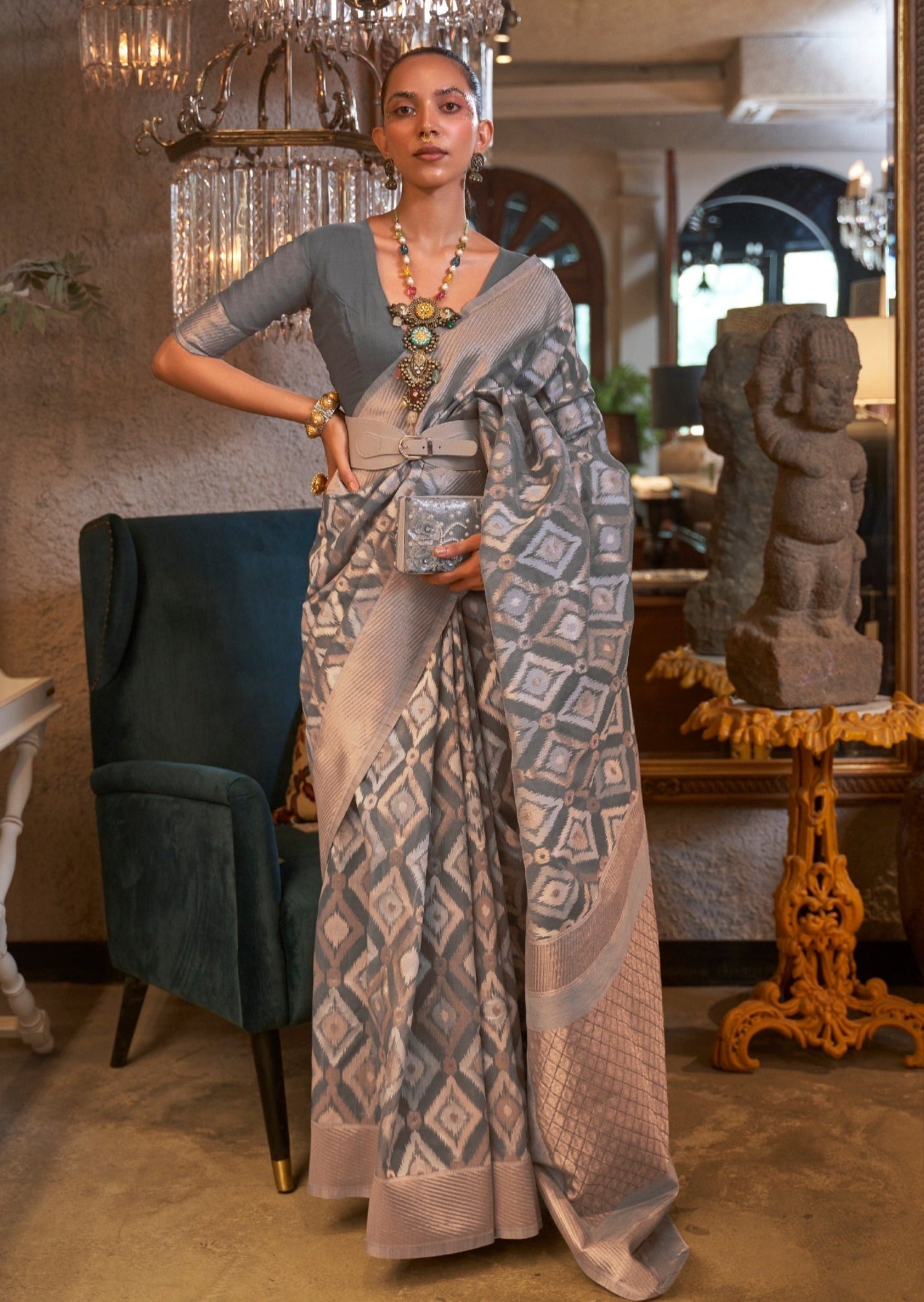 Woman standing in pure-linen-grey-zari work saree with one hand on her waist posing in front of a crystal chandelier and green velvety chair.