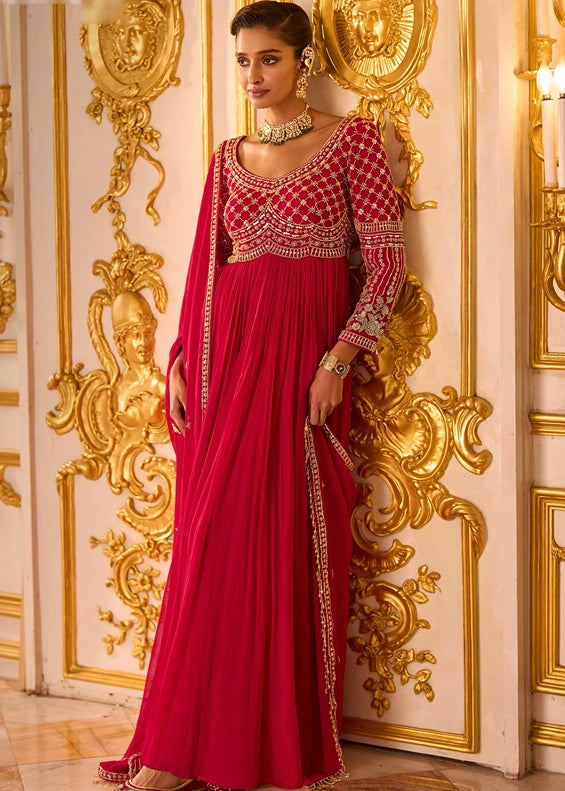 A woman models a traditional Indian anarkali gown in rich red tone with heavy gold embroidery and embellishments, featuring heavily embroidered bodice, long sleeves with a matching net dupatta.