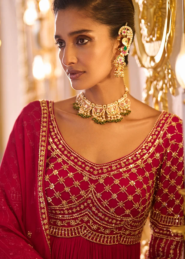 A woman wearing an ornate deep red traditional outfit with extensive gold embroidery, a matching red dupatta, and elaborate gold jewelry with green accents.