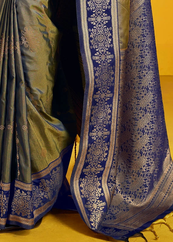 A close-up of a richly patterned kanjivaram silk saree, showing the deep military green color with intricate gold zari floral motifs and border, draped next to a golden yellow background.