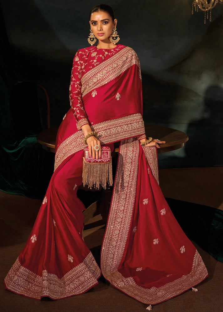 A woman models a royal deep red saree with intricate gold embroidery, paired with a matching embroidered blouse, seated on a wooden table.