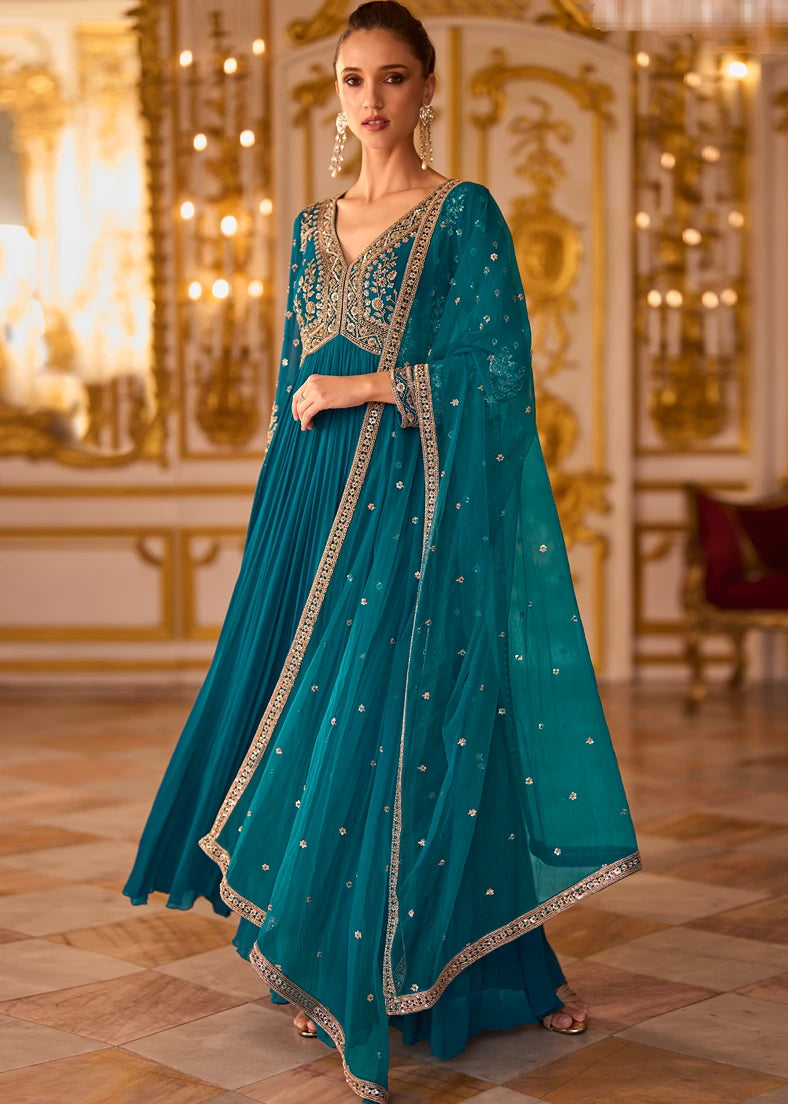 A woman models a traditional teal blue anarkali gown with intricate gold embroidery and a matching dupatta.