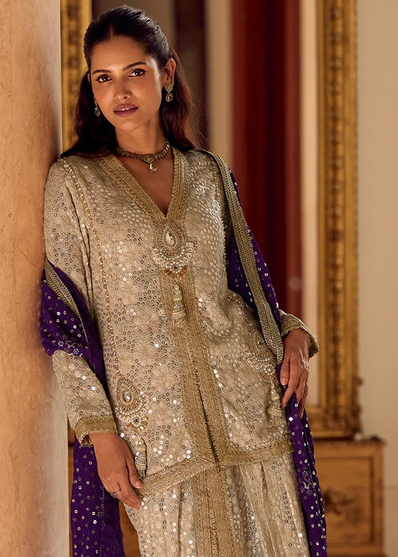A woman models a traditional Indian outfit featuring a heavily embellished beige festive kurta and matching dhoti pants with a deep purple dupatta draped over her shoulder, and indian jewelry.